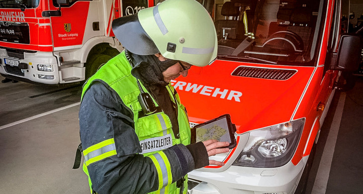 Ein Feuerwehrmann in Uniform und mit Helm steht vor einem Feuerwehrfahrzeug und schaut auf ein Tablet.