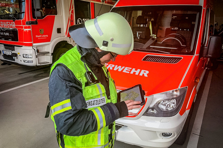 Ein Feuerwehrmann in Uniform und mit Helm steht vor einem Feuerwehrfahrzeug und schaut auf ein Tablet.