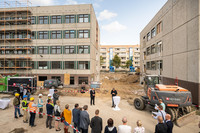 Eine Gruppe von Menschen hat sich vor ehemaligen Schulgebäuden im DDR-Plattenbaustil zu einer Grundsteinlegung versammelt