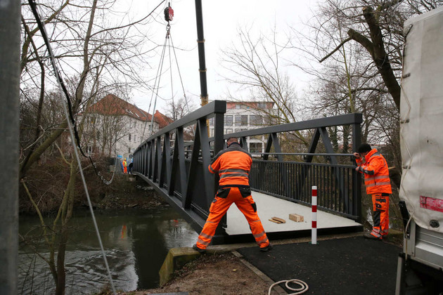 Eine Brücke wird gerade an ihren Platz gesetzt und hängt noch an einem Kran. Zwei Bauarbeiter in oranger Arbeitskleidung justieren sie.