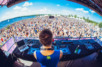 Ein DJ legt auf einer Bühne Musik auf. Vor ihm sind viele Menschen unter freiem Himmel. Das Ufer des Cospudener Sees schließt sich direkt an.