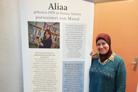 Eine Frau mit Kopftuch steht neben einem Aufsteller mit dem Porträt von "Aliaa"
