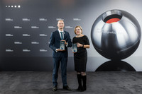 Vor grauem Hintergrund, auf dem auch der Nachhaltigkeitspreis als silberne Kugel abgebildet ist, stehen Herr Trautmann und Frau König und halten je eine Auszeichnung in die Kamera