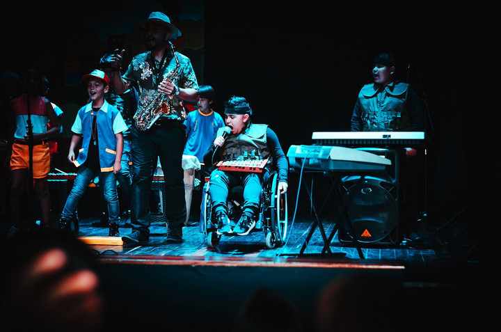 Ein junge im Rollstuhl auf einer Bühne. In der Hand hält er ein Mikrofon. im Hintergrund sind andere Kinder mit Instrumenten.