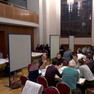 Mehrere Menschengruppen sitzen an verschiedenen Tischen in einem großen Saal und diskutieren.