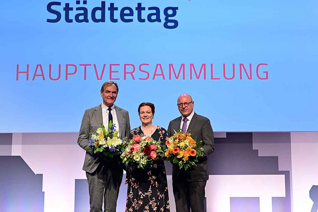 Burkhard Jung, Katja Dörner und Markus Lewe halten Blumensträuße in den Händen, vor einer Leinwand, auf der "Städtetag" steht.