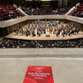 Makroaufnahme rotes Programmheft im Vordergrund, spielendes Orchester im Hintergrund
