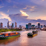 Blick auf einen Fluss, der in blau-violette Töne vom Sonnenuntergang gefärbt ist; auf dem Fluss fahren Containerschiffe, im Hintergrund die Skyline von Ho-Chi-Minh-Stadt mit zahlreichen Hochhäusern
