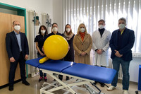 Drei Männer und vier Frauen stehen hinter einer Krankenliege. Eine Frau hält einen gelben Gymnastikball in den Händen.