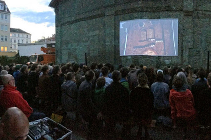 Auf der Außenmauer des ehemaligen Kinos ist eine Leinwand montiert. In der Dämmerung stehen viele Zuschauer davor und schauen einen Film.