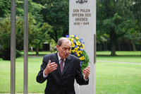 Ein älterer Mann steht gestikulierend vor einem Gedenkstein mit der Aufschrift "Den Polen Opfern des Krieges 1939-1945". Ein Park ist im Hintergrund zu erkennen. 