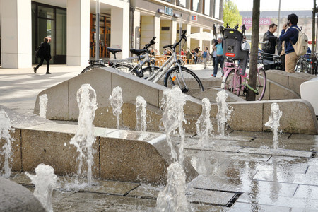 Mehrere kleine Wasserfontänen auf einer Fußgängerzone in Leipzig. Dazwischen sind wellenförmige Steinelemente. Hinter den Fontänen stehen Fahrräder und Leute laufen entlang.