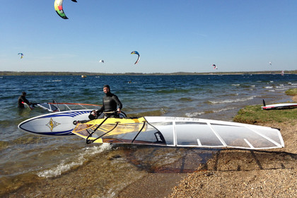 Windsurfer am Strand eines Sees. Im Hintergrund sind viel Segel von Kitesurfern zu sehen.