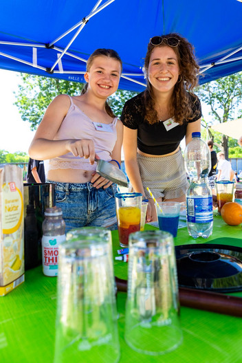 Zwei junge Frauen lächeln in die Kamera. Eine hält eine silberne kleine Schaufel. Vor ihnen steht ein Cocktail.