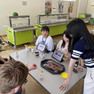 An einem Tisch sitzen auf der einen Seite koreanische Schülerinnen, die über koreanisches Essen sprechen. Gegenüber sitzen deutsche Schüler, die das koreanische Essen auf dem Tisch probieren.