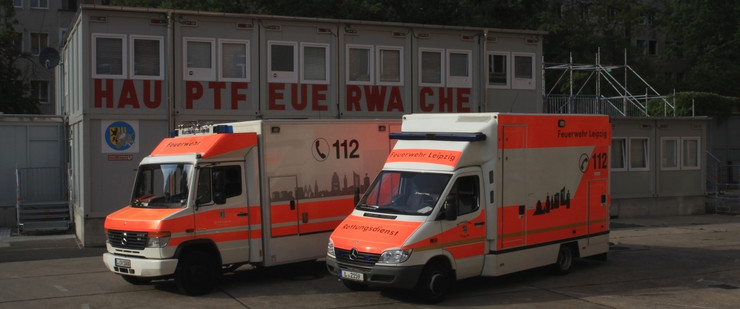Zwei Rettungsdienstfahrzeuge stehen nebeneinander vor einem aus Containern gebauten Gebäude, auf dem in roten Großbuchstaben "Hauptfeuerwache" steht.