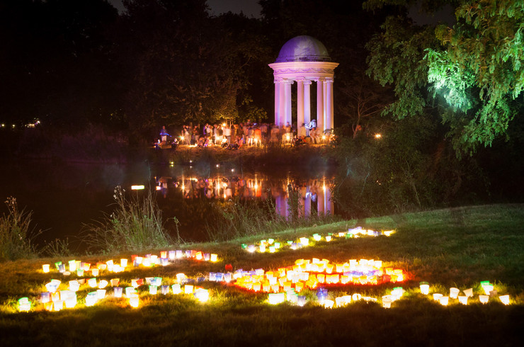 Parkanlage in der Nacht. Ein kleiner Säulentempel ist angeleuchtet. Rund herum sitzen einige Menschen. Eine Wiese vor einem Teich ist mit vielen kleinen Kerzen erleuchtet. 