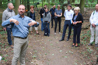 Ein Mann im blauen Hemd mit Brille und lichtem Haar hält im Wald vor rund einem Dutzend Zuhörern einen Vortrag