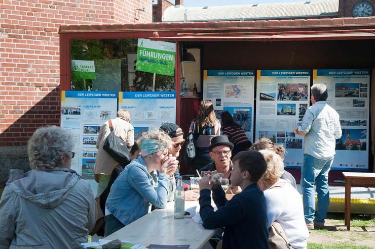Mehrere Personen sitzen an einem Tisch im Freien und essen. Im Hintergrund schauen sich Leute große Infotafeln an.