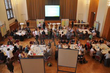 Im Festsaal des Neuen Rathauses sitzen viele Menschen in Gruppen an insgesamt acht Tischen. Sie unterhalten sich und sammeln Ideen auf Karten an einer Pinnwand.