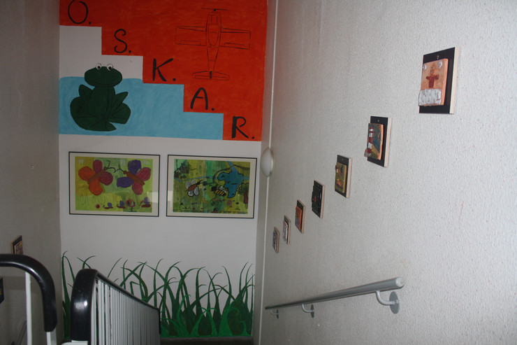 Der Treppenaufgang ist mit einem Frosch und dem Schriftzug OSKAR bemalt. Darunter hängen zwei von Kindern gestaltete Bilder. Über dem Treppengeländer sind Keramiken in Bildform aufgehängt.