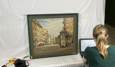 Eine Frau sitzt mit dem Rücken zur Kamera gewandt vor einem Laptop und betrachtet ein Gemälde.
