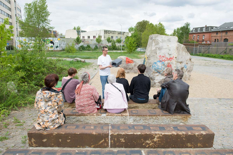 Eine Menschengruppe sitzt auf Sitzmöglichkeiten auf dem Gelände des ehemaligen Plagwitzer Güterbahnhofes. Vor der Gruppe steht ein Mann und redet.