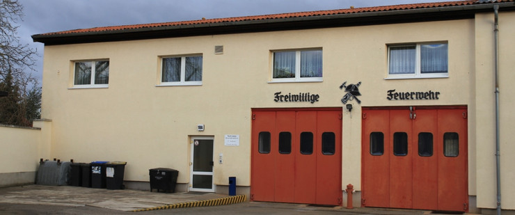 Ein gelblich gestrichenes Haus. Rechts zwei große, rote Tore, über denen "Freiwillige Feuerwehr" geschrieben steht. In der Mitte eine Eingastür. Die erste Etage besitzt vier Fenster.