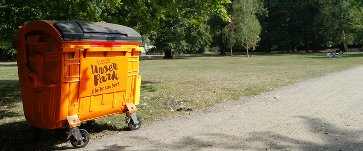 Vor einer gepflegten und aufgeräumten Parkwiese steht ein großer oranger Müllcontainer mit dem aufgedruckten Logo "Unser Park".