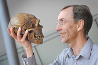 Ein älterer Herr mit Brille hält sich mit einer Hand den Schädel eines Neandertalers vors Gesicht und betrachtet ihn lächelnd.