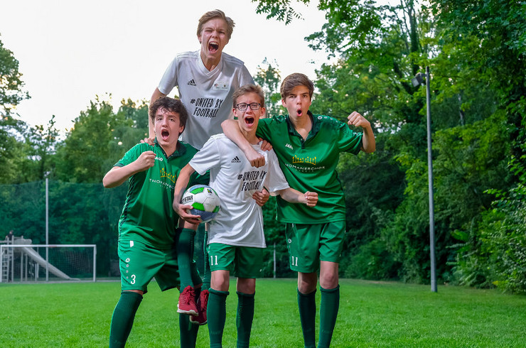 Vier Jungen in Fußballtrikots und mit einem Ball in der Hand stehen zusammen, schreien und feuern sich an.
