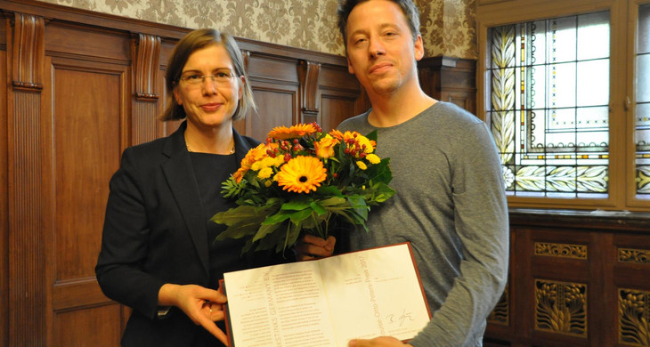 Die Bürgermeisterin Dr. Skadi Jennicke und Nils Pickert von Pinkstinks Germany posieren mit einem Blumenstrauß und er Urkunde zum Louise-Otto-Peters-Preis 2017 für ein Foto. Sie lachen in die Kamera.