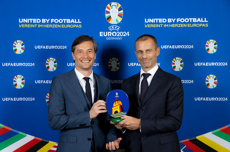Sportbürgermeister Heiko Rosenthal und UEFA-Präsident Aleksander Čeferin halten gemeinsam eine Plakete in der Hand, die ein stilisiertes Völkerschlachtdenkmal zeigt.