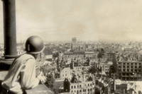 Ein Mann mit Helm steht am Geländer und schaut von einem Turm auf die zerstörte Stadt. Vielen Häusern fehlt das Dach.