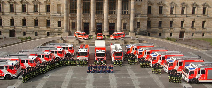 Im Halbkreis aufgestellte Einsatzfahrzeuge von Berufs- und Freiwilliger Feuerwehr nebst Sondertechnik mit davor angetretenen Feuerwehrangehörigen