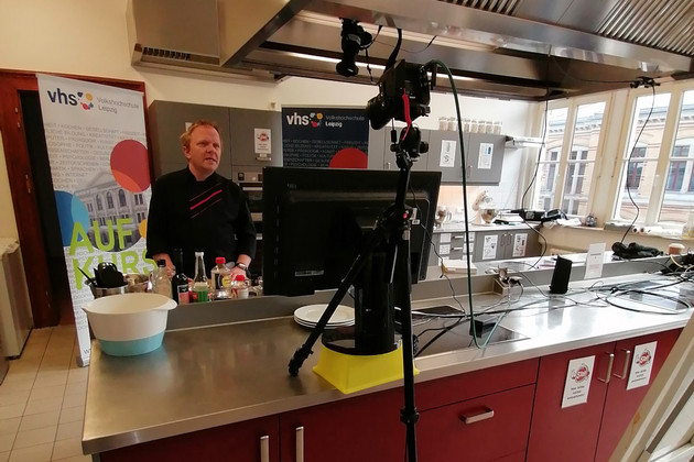 Ein Mann steht in einer großen Küche, vor ihm steht eine Kamera auf einem Stativ.