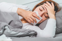 Frau im Bett mit Taschentuch vor der Nase und einer Hand auf dem Kopf.