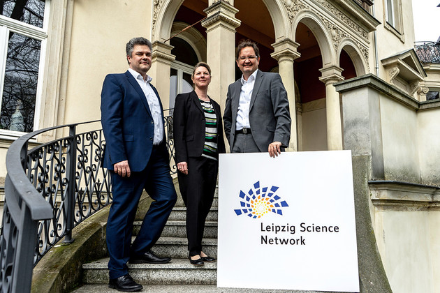 Drei Mitglieder des Vorstands stehen auf einer steinernen Treppe. Der Mann ganz rechts hat ein bedrucktes Schild in der Hand. Auf dem Schild steht "Leipzig Science Network" mit dem Logo daneben.