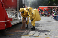 Bei einer Vorführung schütten zwei Feuerwehrmänner mit gelben Chemikalienschutzanzügen Bindemittel auf eine Flüssigkeit am Boden. Im Hintergrund beobachten zahlreiche Zuschauer das Geschehen.