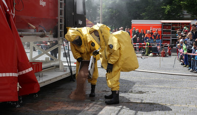 Bei einer Vorführung schütten zwei Feuerwehrmänner mit gelben Chemikalienschutzanzügen Bindemittel auf eine Flüssigkeit am Boden. Im Hintergrund beobachten zahlreiche Zuschauer das Geschehen.