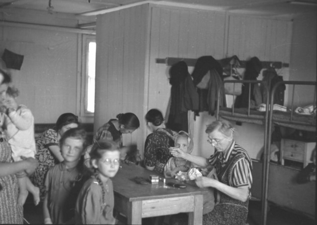 Frauen und Kinder an einem Tisch im Innenraum der Baracke des Umsiedlungslagers mit Doppelstockbett im Hintergrund