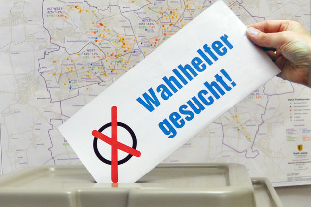 Hand steckt Zettel mit Aufschrift "Wahlhelfer gesucht" in Wahlurne