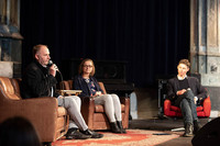 Zwei Männer und eine Frau sitzen auf braunen und roten Sesseln im Halbkreis auf einer Bühne. Einer spricht in ein Mikrofon.