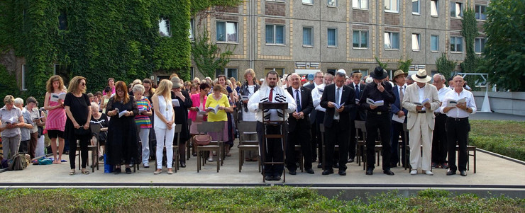 Eine große Menschengruppe betet stehend gemeinsam mit dem Rabbi in der vordersten Reihe, die Herren rechts im Bild, die Frauen links im Bild an der Gedenkstätte