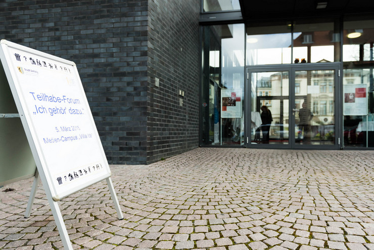 Vor einem Haus mit einer Fassade aus schwarzen Ziegeln und mit einem großen Glaseingang steht ein Aufsteller mit der Aufschrift "Teilhabe-Forum, Ich gehör' dazu, 5. März 2015, Medien-Campus Villa Ida".