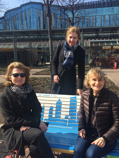 Zwei Damen der Lyoner Delegation, die zur Buchmesse 2017 in Leipzig zu Besuch war, sitzen am Augustusplatz auf der Partnerstadtbank Lyons. Dahinter steht Frau Dr. Marusch-Krohn vom Referat Internationale Zusammenarbeit der Stadt Leipzig.