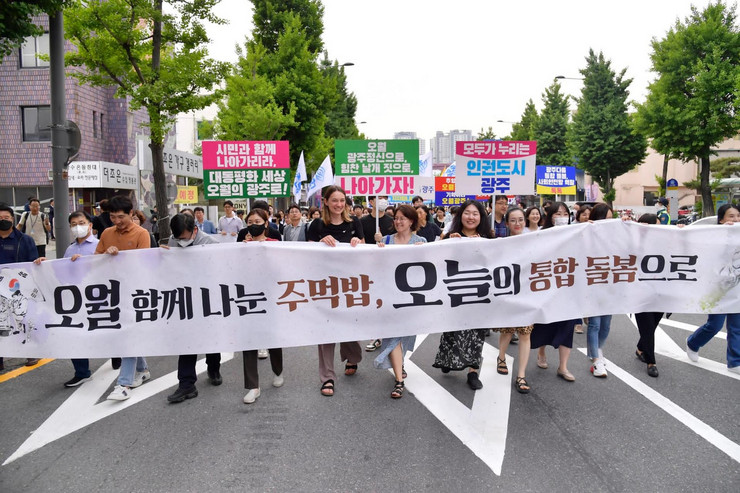Auf einer Straße läuft eine große Gruppe von Personen. Die Personen in der ersten Reihe halten einen langen Banner in den Händen mit einem Schriftzug in koreanischer Sprache..