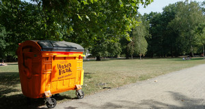 Vor einer gepflegten und aufgeräumten Parkwiese steht ein großer oranger Müllcontainer mit dem aufgedruckten Logo "Unser Park".