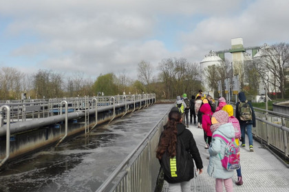 Kinder laufen über eine Brücke, links von ihnen ist ein großes Becken mit Wasser gefüllt. Im Hintergrund sieht man eine große Kläranlage.