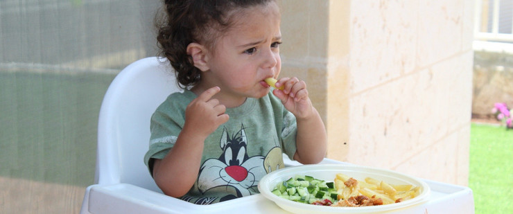 Ein kleines Mädchen sitzt im Hochstuhl und steckt sich eine Pommes in den Mund. Auf dem Tischchen vor ihr steht ein Teller mit Kartoffelstreifen und Gemüse.